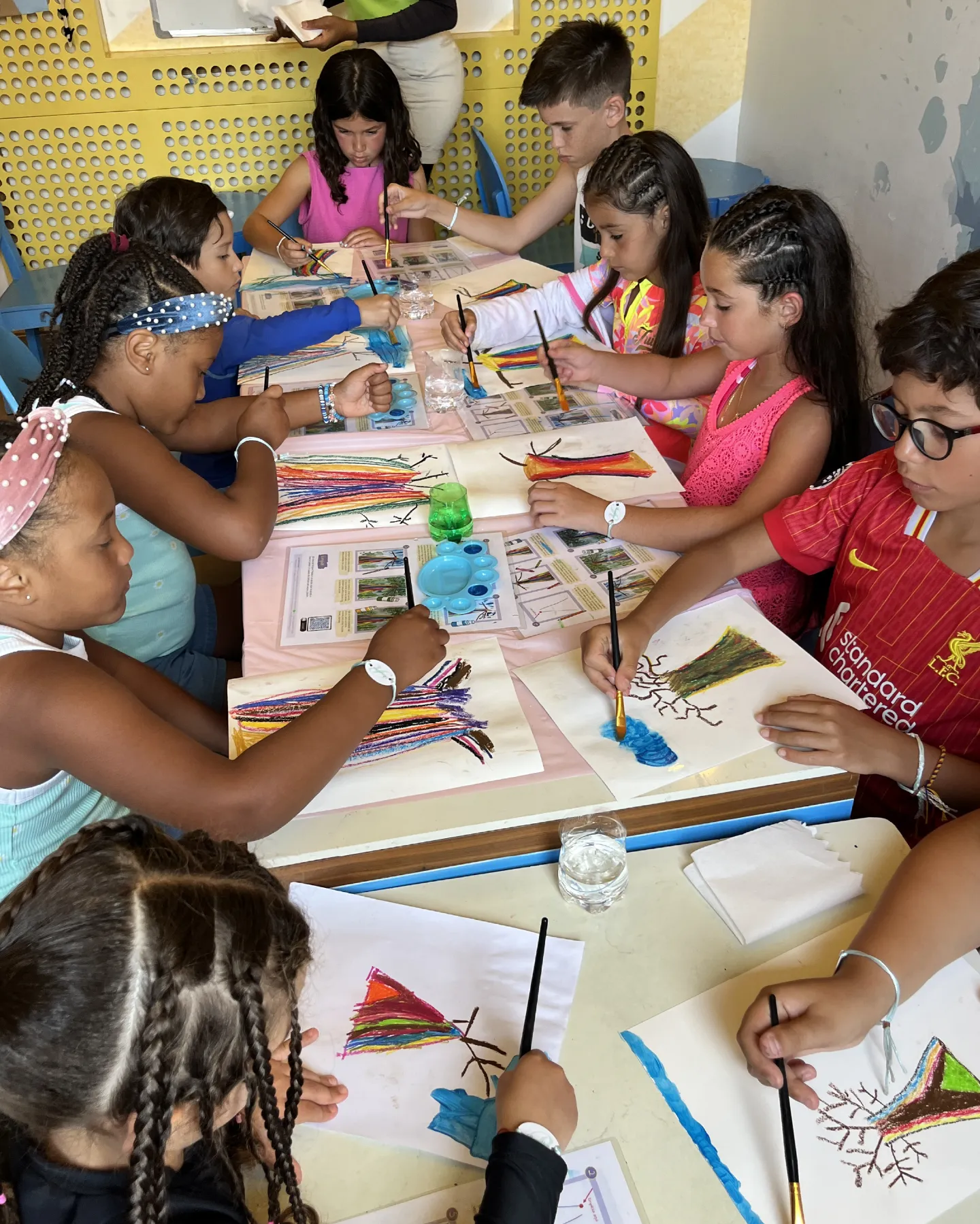 A lively group of diverse children with braided hairstyles, including girls in pink, teal, and floral tops and a boy in a red Liverpool soccer jersey, sit closely around a pink-clothed table in a sunny yellow-walled room with blue chairs, deeply focused on a collaborative art project. They weave rainbow-colored paper strips into tree branches and roots on white sheets, paint with blue and green brushes, and refer to instructional worksheets featuring tree illustrations. Scattered supplies include glue sticks, water cups, palettes, and yarn. An adult woman in green oversees from the side, fostering creativity and joy in this vibrant workshop scene.