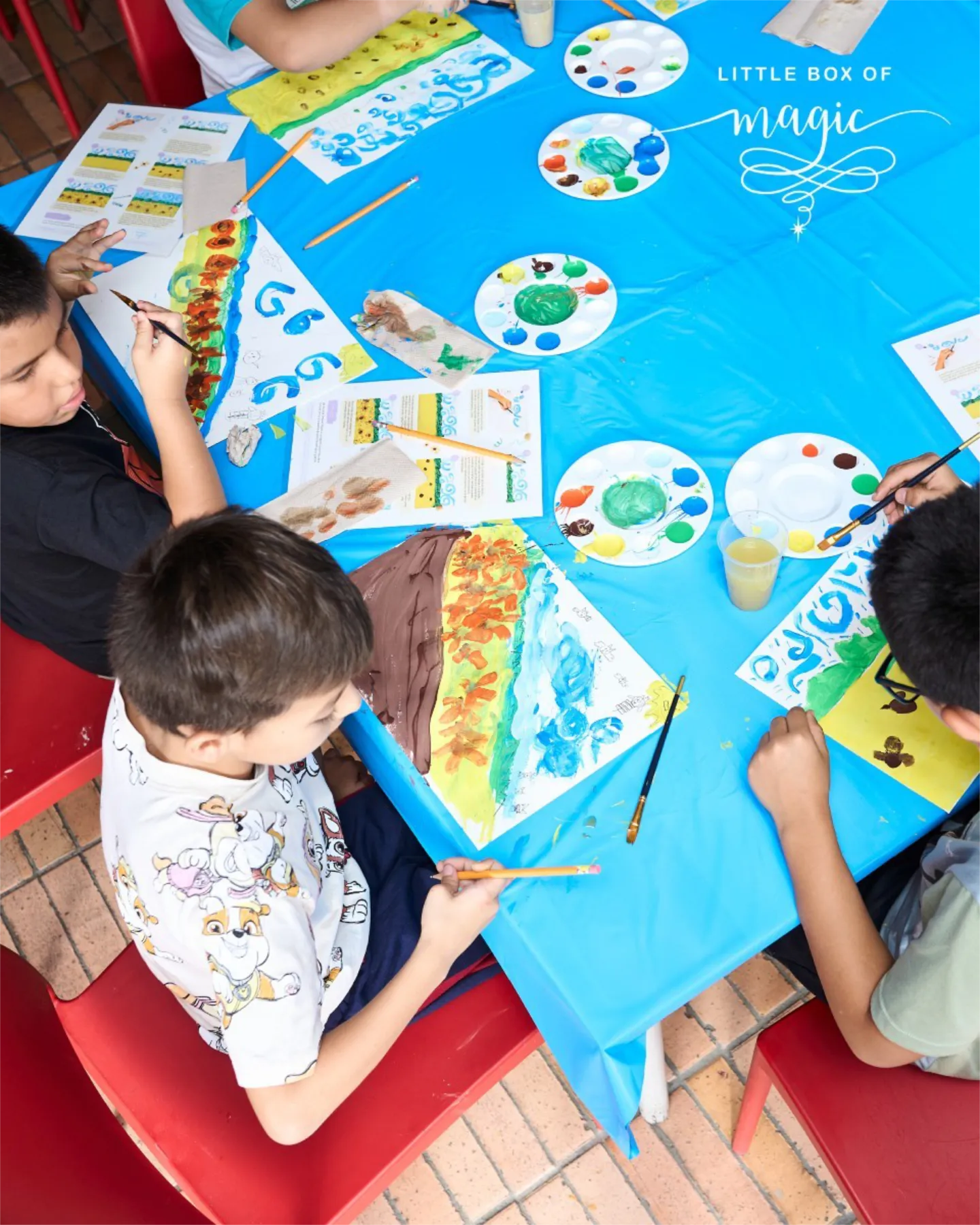 A joyful group of three young boys with short dark hair, dressed in casual T-shirts and shorts, huddle around a blue-covered table outdoors on a tiled patio, deeply engaged in painting. The boy on the left, in a black T-shirt, dips a brush into orange paint on a palette; the center boy, in a white T-shirt with cartoon dog prints and blue shorts, holds a pencil thoughtfully; the right boy, in a light green T-shirt, paints with a blue brush. Their white canvases feature abstract landscapes with rainbow stripes, swirling blues and greens, and yellow accents. Scattered supplies include colorful paint palettes, brushes, pencils, worksheets with instructions, cardboard cutouts, and a yellow juice cup. Red chairs frame the scene, with elegant white script "Little Box of Magic" and swirling flourishes at the top.