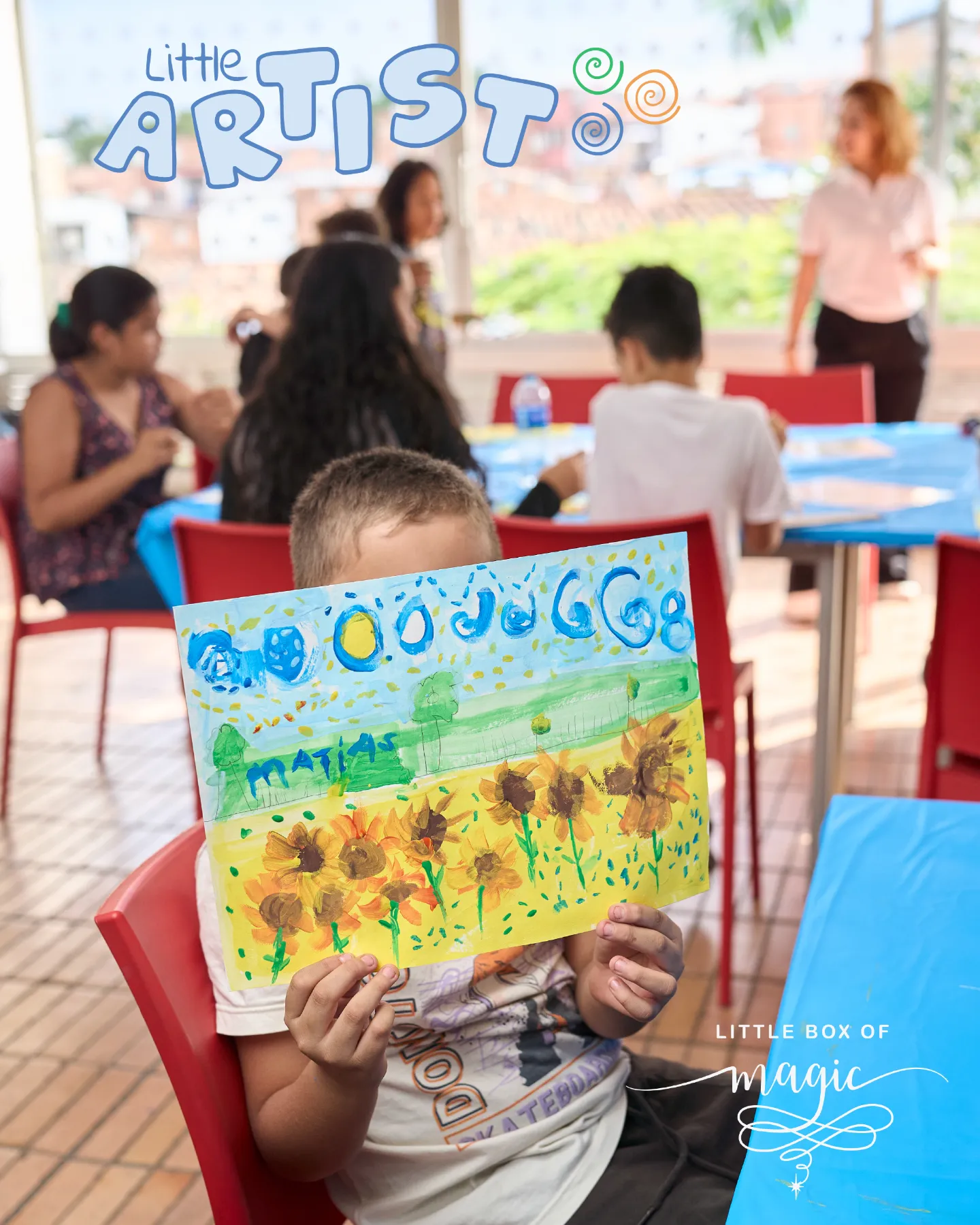 A beaming young boy with short brown hair, wearing a white T-shirt with colorful prints and khaki shorts, sits in a red chair holding up his vibrant watercolor painting of a sunny yellow field of orange sunflowers under a blue sky dotted with white clouds, green trees in the distance, and a large yellow sun, signed "MATIAS" at the bottom. In the softly blurred background, a group of diverse adults and children engage in an art session around blue-covered tables and red chairs in a sunlit room with large windows overlooking palm trees and brick walls. Blue bubbly text overhead reads "Little Artist" with green and orange swirls, and at the bottom, the elegant "Little Box of Magic" logo with decorative flourishes and a star.